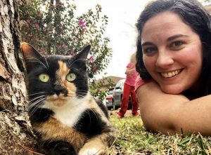 Ground level selfie of a calico cat and a woman lying next to her, smiling