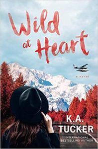 A photorealistic book cover with a woman facing away from the camera, looking at an empty road, fiery red trees in the process of changing colors, and a snowy mountain in the distance. A plan flies in the sky, and she's holding onto her black hat with one hand. The title is in red brushstroke font in the sky: Wild at Heart. The author's name appears over the road: K.A. Tucker. 